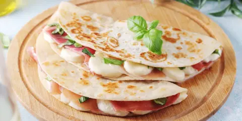 Due piadine farcite con prosciutto crudo, mozzarella e basilico, servite su un tagliere di legno.