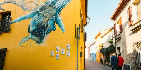 Un murale di balene blu su un muro giallo a San Giuliano Rimini. Una coppia passeggia lungo la strada acciottolata.