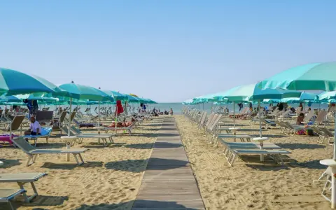 Una passerella di legno conduce alla spiaggia di San Giuliano Mare, fiancheggiata da file di lettini e ombrelloni azzurri sotto un cielo limpido.