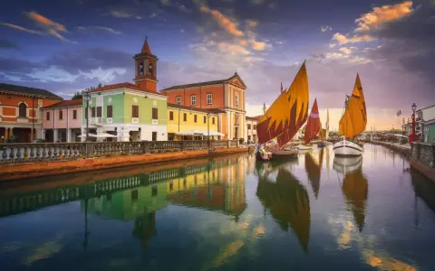 Navi a vela colorate navigano sul canale di Cesenatico al tramonto, con edifici vivaci e riflessi sull'acqua. Meglio Cervia o Cesenatico? Cesenatico offre questo spettacolo.