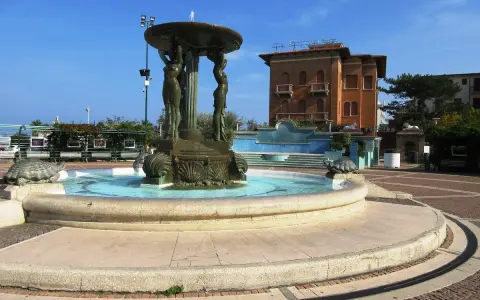 Fontana con statue di donne e tartarughe in piazza Primo Maggio Cattolica, con un edificio in mattoni sullo sfondo.