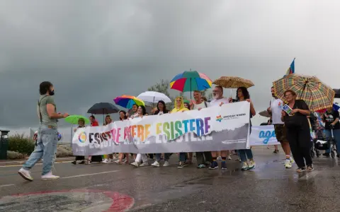 Persone con ombrelli colorati sfilano a Rimini Pride, tenendo uno striscione arcobaleno con scritto "Esistere Mere Esistere".
