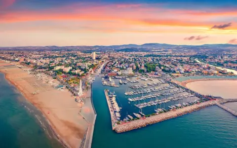 Tramonto sul lungomare di Rimini, con la spiaggia dorata, il porto turistico pieno di barche e la città che si estende fino alle colline.