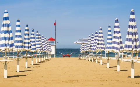 Filetto di ombrelloni a righe blu e bianche sulla spiaggia di Rivabella di Rimini, con mare calmo e cielo azzurro.