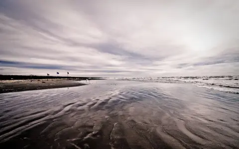 Spiaggia deserta con onde che lambiscono la riva, tre bandierine piantate nella sabbia bagnata. Un'atmosfera malinconica per questo mare d'inverno.