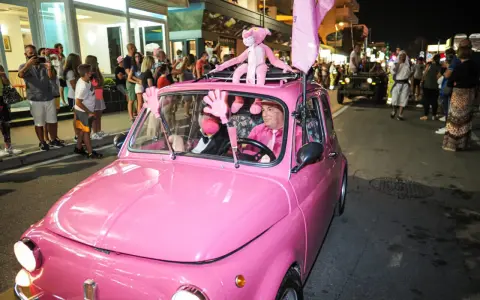 Un'auto rosa shocking sfila a Rimini durante la Notte Rosa, con un pupazzo a forma di pantera rosa sul tetto e un uomo al volante vestito a tema.
