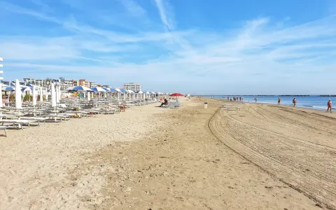 Spiaggia di Ravenna Lido Adriano con file di ombrelloni e lettini, bagnanti che passeggiano sul bagnasciuga sotto un cielo azzurro.