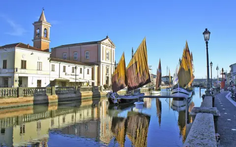 Traditional sailboats with colorful sails line a canal in Cesenatico, Italy, with buildings and a church reflected in the water.