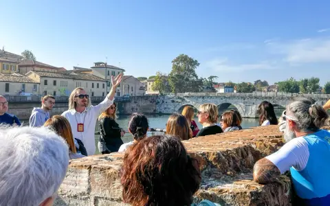 Guida turistica con gruppo a Rimini, che ascolta storie sui **eventi rimini** e la storia locale, con un ponte antico sullo sfondo.