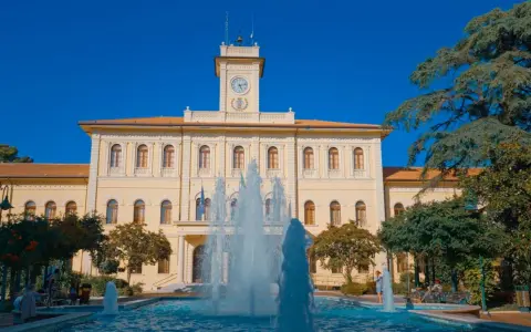 Fontana zampillante nella piazza Roosevelt di Cattolica, con un edificio giallo e un cielo azzurro.