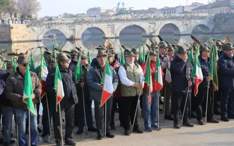 Uomini con cappelli alpini e bandiere italiane partecipano all'adunata alpini a Rimini, con un ponte storico sullo sfondo.