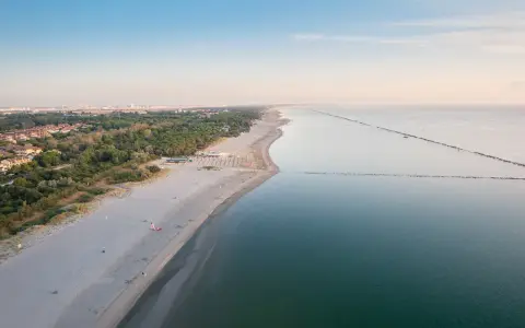 Vasta spiaggia dorata e mare calmo, con file di ombrelloni che delineano un lido di classe. All'orizzonte, la costa punteggiata di verde e costruzioni.