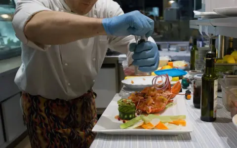 Chef con guanti blu macina pepe su un'aragosta, un piatto che sembra uscito da "lo zodiaco Rimini".