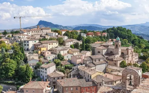 Panoramica di un borgo collinare con tetti rossi e una chiesa con campanile, incorniciato dal verde e dalle montagne. Un paesaggio che evoca la bellezza di Rimini e dintorni.