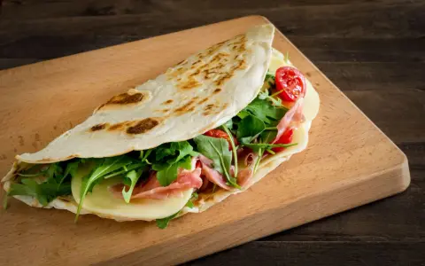 Piadina romagnola, un classico dal sapore autentico, ripiena di prosciutto, formaggio, rucola e pomodorini.
