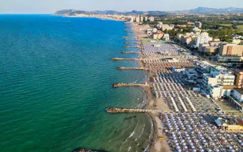 Vista aerea della costa adriatica, con la spiaggia affollata di ombrelloni e stabilimenti balneari, che si estende fino a Cattolica e Misano.