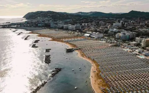 Vista aerea della cattolica spiaggia, con file ordinate di ombrelloni e lettini che si estendono lungo la costa, mentre il mare luccica sotto il sole.
