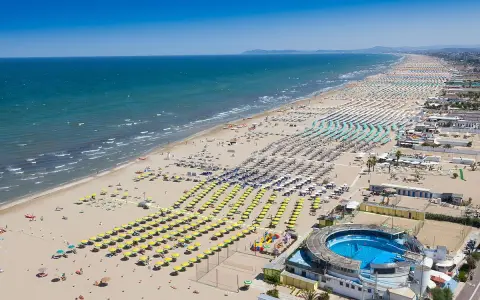 La riviera di Rimini in pieno sole, con un mare azzurro e una spiaggia affollatissima di ombrelloni colorati e bagnanti.
