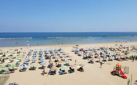 Spiaggia libera Viserba affollata di ombrelloni e lettini sotto un cielo azzurro. Bambini giocano nell'area attrezzata.