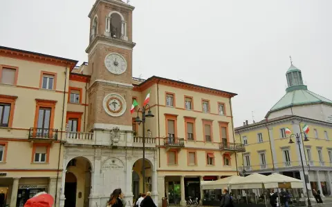 La piazza Rimini, con il suo imponente campanile e gli edifici storici, accoglie visitatori e residenti sotto un cielo nuvoloso.