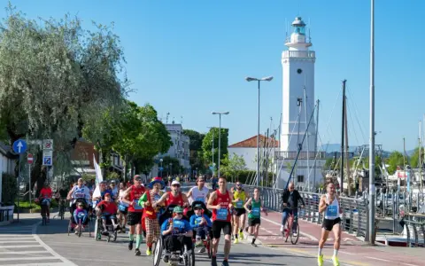 Atleti corrono lungo il percorso della Rimini Marathon, con il faro bianco e il porto sullo sfondo.