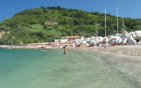 Spiaggia affollata con gente che nuota e prende il sole. Un promontorio verde domina la scena. Meglio Riccione o Gabicce Mare? Difficile scegliere!