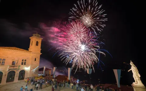 Spettacolo pirotecnico illumina il cielo notturno per la festa di San Marino. Folla ammira i fuochi d'artificio da una piazza con un edificio storico.