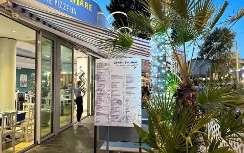 Osteria del Mare, ristorante pizzeria a Rimini centro, con menù esposto e tavoli all'esterno. Palme illuminate con lucine.