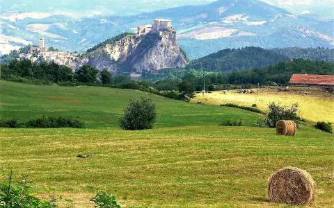 Campagna con borghi vicino Rimini, colline verdi, balle di fieno e un castello arroccato su uno sperone roccioso.