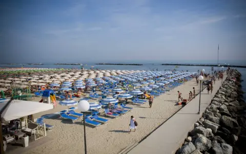Gatteo Mare lungomare affollato di ombrelloni e lettini, con persone che passeggiano sul molo roccioso e sulla sabbia.