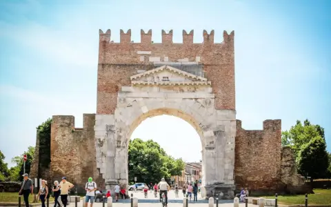 L'imponente arco d'augusto rimini, con le sue antiche pietre e la torre in mattoni, fa da sfondo a una vivace piazza con persone che passeggiano e ciclisti.