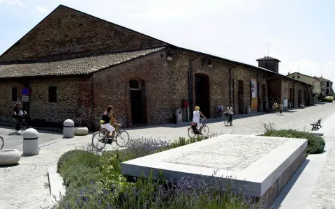Passeggiata in bicicletta davanti ai Magazzini del Sale di Cervia, con persone che pedalano su un lungomare acciottolato, tra aiuole fiorite e antichi edifici in mattoni.