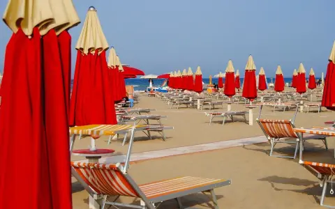 File di ombrelloni rossi e lettini prendisole su una spiaggia, forse a Riccione o Rimini, in attesa dei bagnanti.