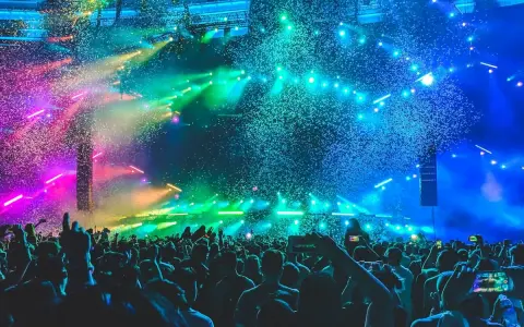 Folla in delirio a un concerto a Rimini, con coriandoli colorati e luci psichedeliche che illuminano la scena.