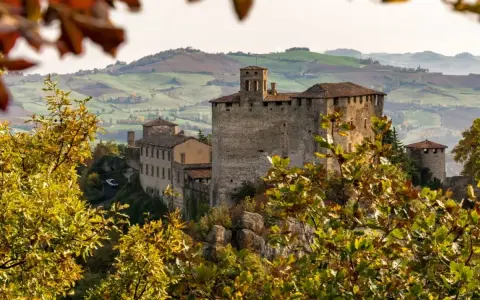 Castello medievale su una collina, circondato da alberi con foglie autunnali dai colori caldi. Il paesaggio collinare sullo sfondo evoca un senso di pace in questo mare autunno.