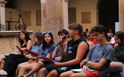 Giovani ascoltano un discorso, alcuni con fogli rossi in mano, durante un evento culturale a Rimini. Un ragazzo tiene un microfono.
