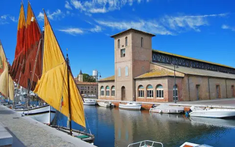 Vele colorate di barche tradizionali ormeggiate nel porto canale di Cervia mare, con l'antico edificio delle saline sullo sfondo.