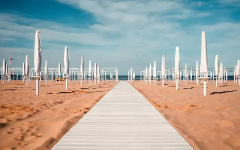 Passerella bianca su sabbia dorata, file di ombrelloni chiusi e strutture bianche sotto un cielo azzurro. Un'immagine serena del mare in Emilia-Romagna.