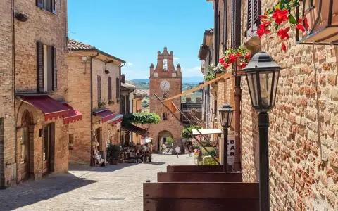 Un vicolo acciottolato a Gradara, con edifici in mattoni, fiori rossi e un campanile con orologio. Perfetto per scoprire cosa vedere.