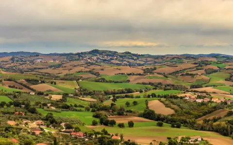 Dolci colline verdi e marroni della Valconca, Rimini, punteggiate da case e alberi, sotto un cielo nuvoloso.