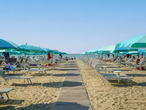 Una passerella di legno conduce alla spiaggia di San Giuliano Mare, fiancheggiata da file di lettini e ombrelloni azzurri sotto un cielo limpido.