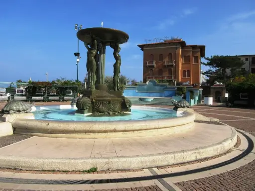 Fontana con statue di donne e tartarughe in piazza Primo Maggio Cattolica, con un edificio in mattoni sullo sfondo.