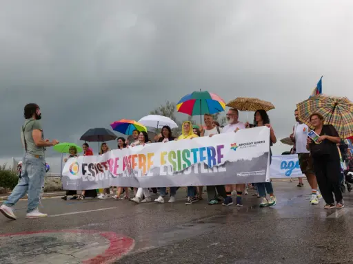 Persone con ombrelli colorati sfilano a Rimini Pride, tenendo uno striscione arcobaleno con scritto "Esistere Mere Esistere".