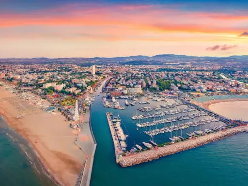 Tramonto sul lungomare di Rimini, con la spiaggia dorata, il porto turistico pieno di barche e la città che si estende fino alle colline.