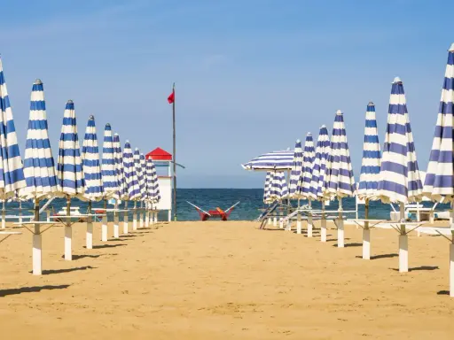 Filetto di ombrelloni a righe blu e bianche sulla spiaggia di Rivabella di Rimini, con mare calmo e cielo azzurro.