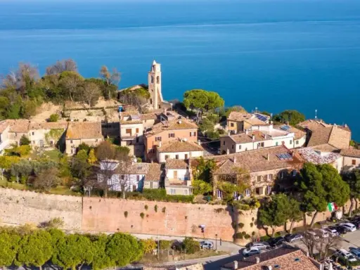Borgo antico con campanile affacciato sul mare Adriatico. Il centro storico del lungomare Gabicce Mare, con tetti rossi e vegetazione rigogliosa, si estende verso l'azzurro intenso dell'acqua.