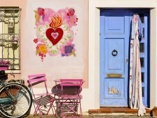 Un angolo pittoresco di Borgo San Giuliano a Rimini: una porta blu, un cuore fiammeggiante dipinto, una bicicletta e sedie rosa.