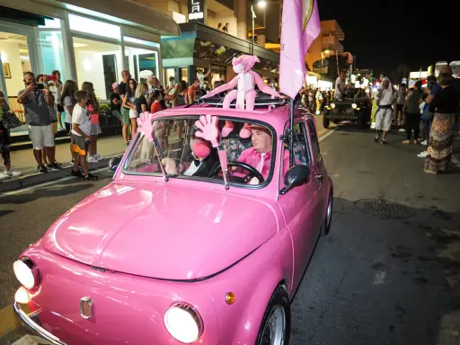 Un'auto rosa shocking sfila a Rimini durante la Notte Rosa, con un pupazzo a forma di pantera rosa sul tetto e un uomo al volante vestito a tema.