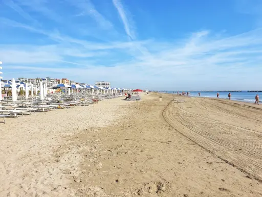 Spiaggia di Ravenna Lido Adriano con file di ombrelloni e lettini, bagnanti che passeggiano sul bagnasciuga sotto un cielo azzurro.