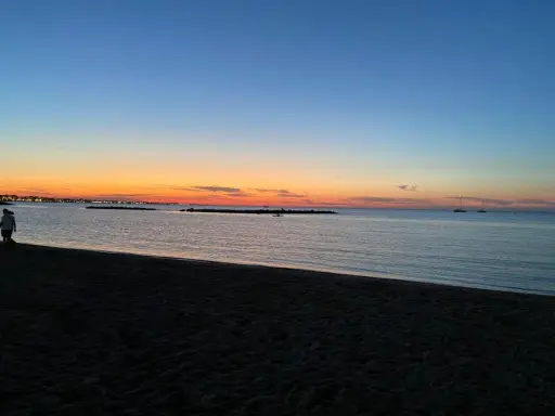 Tramonto sulla spiaggia di Rimini, con il cielo che sfuma dall'arancione al blu. Due persone passeggiano sulla sabbia scura, mentre in mare si vedono barche a vela.